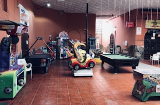 An indoor arcade area featuring various gaming machines, including an air hockey table and a ride-on toy car. The room has a tiled floor and several colorful gaming devices scattered around. Chairs and arcade signage are visible, creating a playful and entertainment-focused atmosphere.