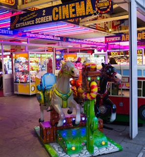 A vibrant amusement arcade featuring various games and bright neon signs. Mechanical horse rides are in the foreground, surrounded by claw machines with plush toys. The entrance is adorned with signs for a cinema and arcade, creating a lively and colorful atmosphere.