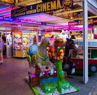 A vibrant amusement arcade featuring various games and bright neon signs. Mechanical horse rides are in the foreground, surrounded by claw machines with plush toys. The entrance is adorned with signs for a cinema and arcade, creating a lively and colorful atmosphere.